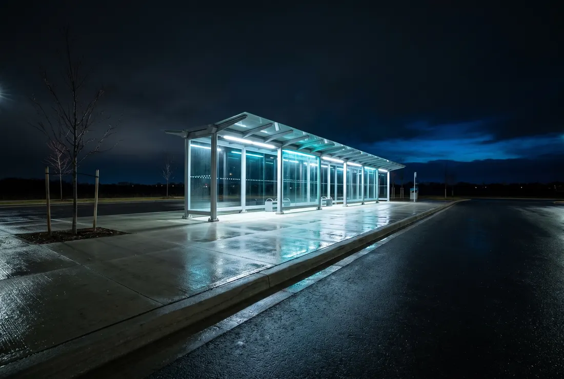Scène générée : un abri de transport en commun vide sous une longue marquise de verre éclairée en cyan la nuit, un quai mouillé et un ciel sombre.