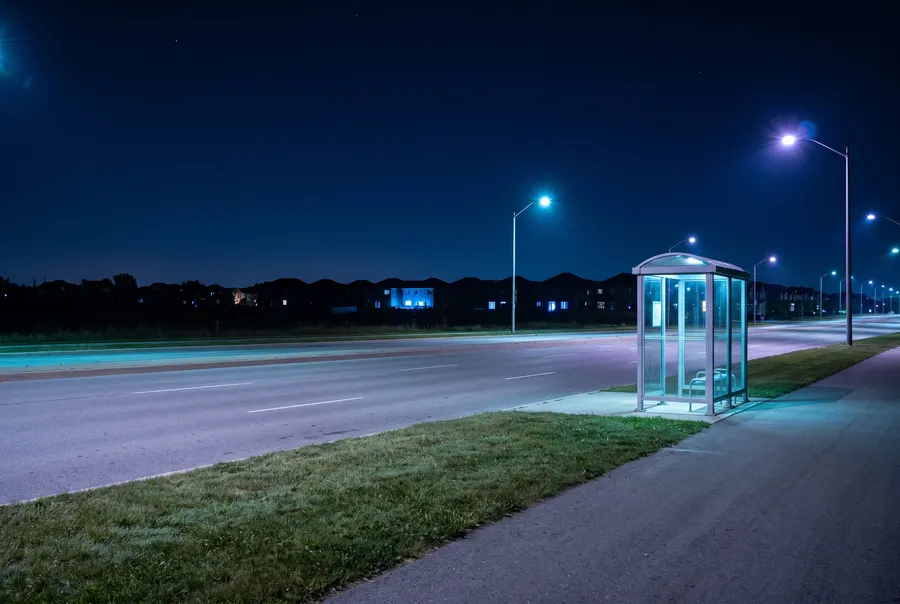 Generated scene: a single glass bus shelter beside a wide empty road in the small hours, lit by blue-white streetlights.